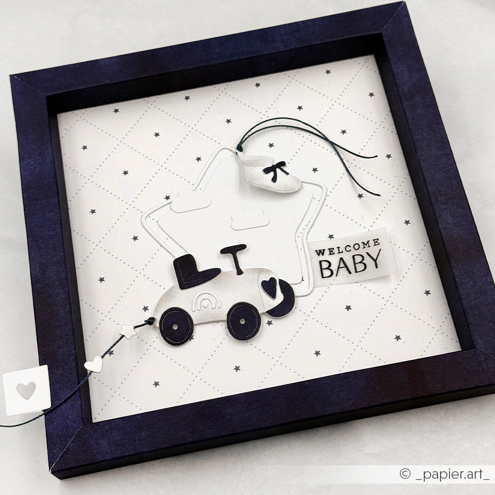 Gerahmtes Papierkunstwerk eines Kinderwagens mit "Welcome Baby"-Anhänger, Herz-Akzenten und lilafarbenem Rand, hergestellt mit Alexandra Renke Designpapieren und der Motivstanze 5 Sternrahmen für zusätzlichen Charme.