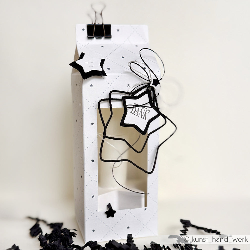 Weiße Geschenkbox mit Sternausschnitten aus der Alexandra Renke Motivstanze 5 Sternrahmen, veredelt mit schwarzen Bändern und einem kleinen "DANK"-Anhänger.