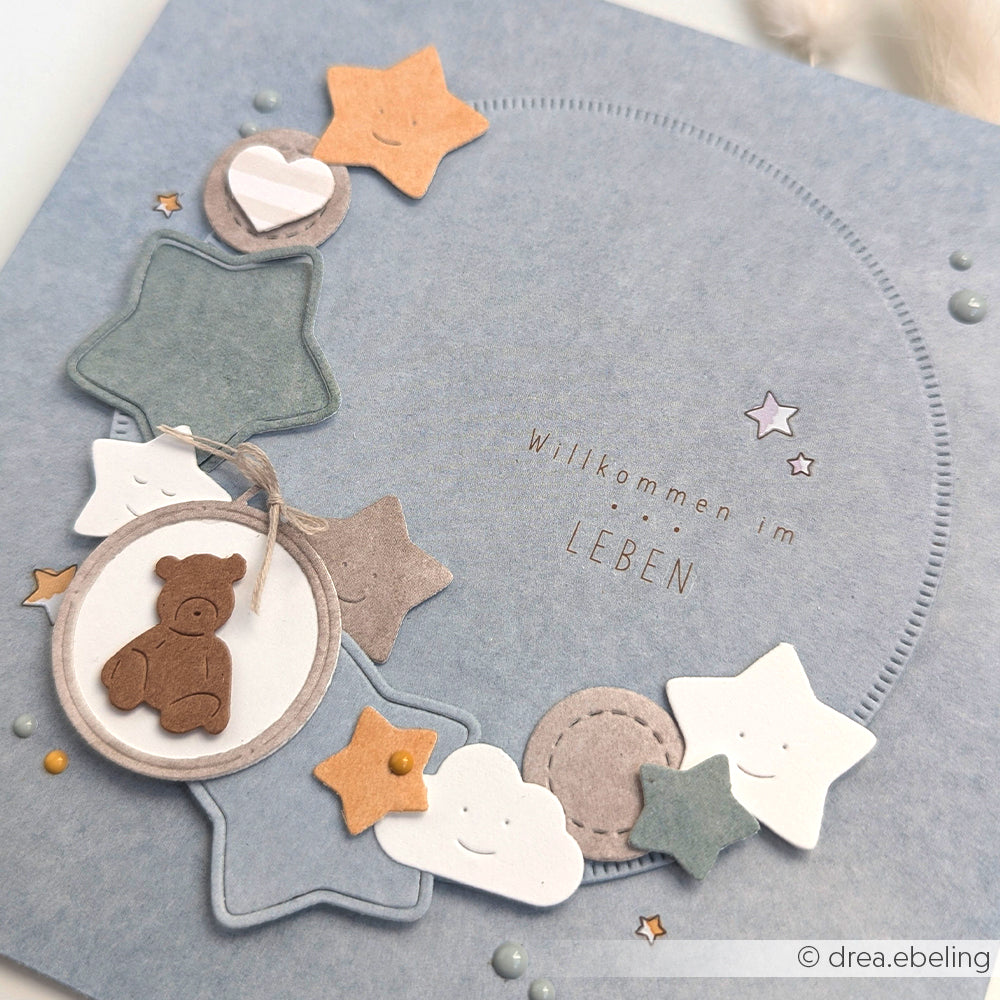 Blaue Karte von Alexandra Renke mit Sternen, Wolken, einem Teddybär-Anhänger und "Willkommen im Leben" - hergestellt mit Designpapieren und der Motivstanze 3 Sterne DieCut Systeme für detaillierte Akzente.