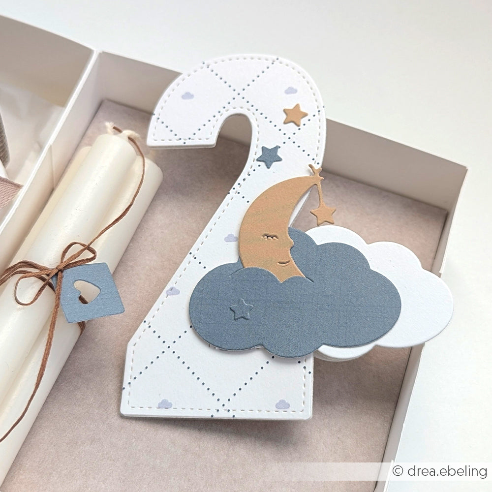 Eine Karte in Form einer Nummer 2 aus eleganten Alexandra Renke Designpapieren, verziert mit Wolken, einer Mondsichel und Sternen, steht neben einer gebundenen Kerze.