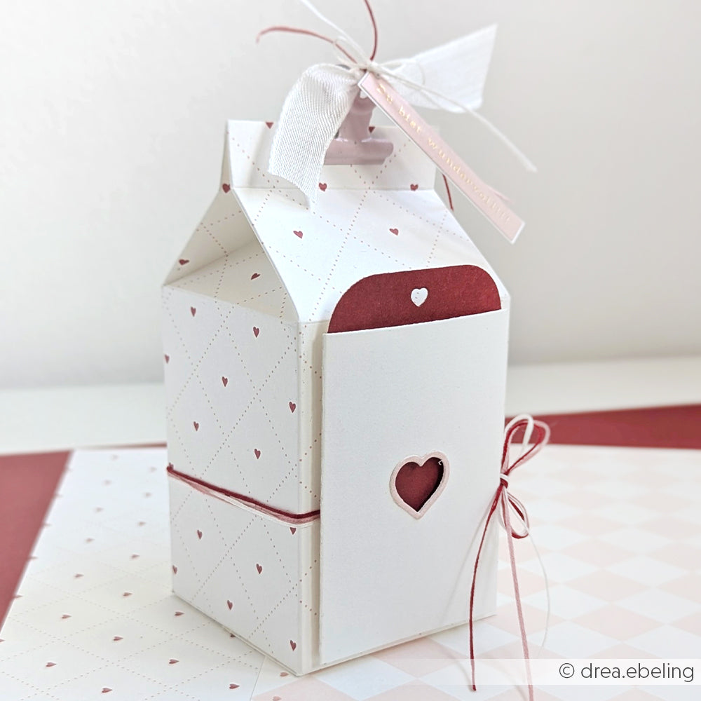 Weiße, herzförmige Geschenkbox mit Herzausschnitt, hergestellt mit der Motivstanze Etikett im Tütchen-Herz von Alexandra Renke und eleganten 200g Designpapieren, veredelt mit Band und Schleife auf passendem Hintergrund.