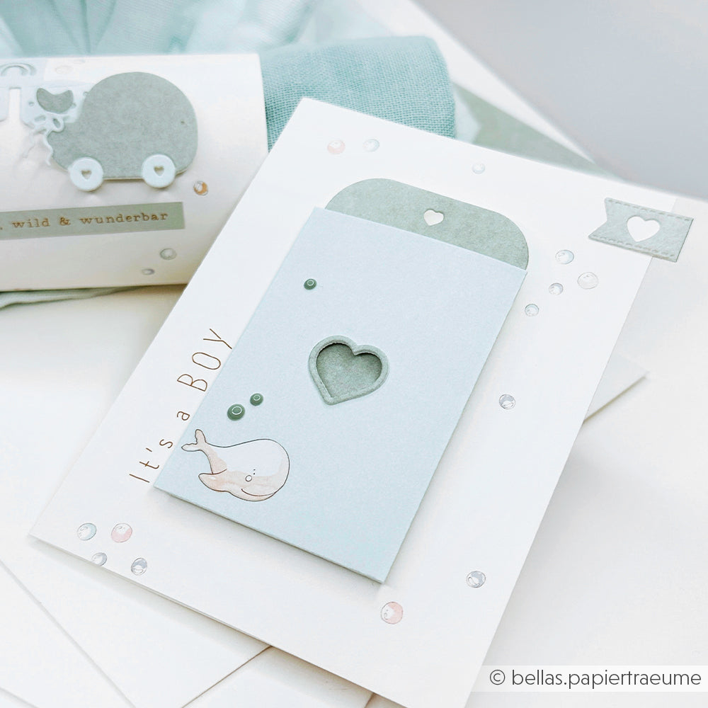 Hellblaue Babyparty-Karte von Alexandra Renke mit "It's a BOY", einem Wal, Herzausschnitten aus Stanzen für Designpapiere und pastellfarbenen Verzierungen.