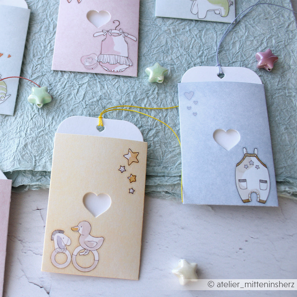 Zwei Geschenkanhänger aus pastellfarbenem Papier mit Herzausschnitten, die mit den Schneidewerkzeugen von Alexandra Renke hergestellt wurden, zeigen Illustrationen zum Thema Sterne, Spielzeug und Babys.
