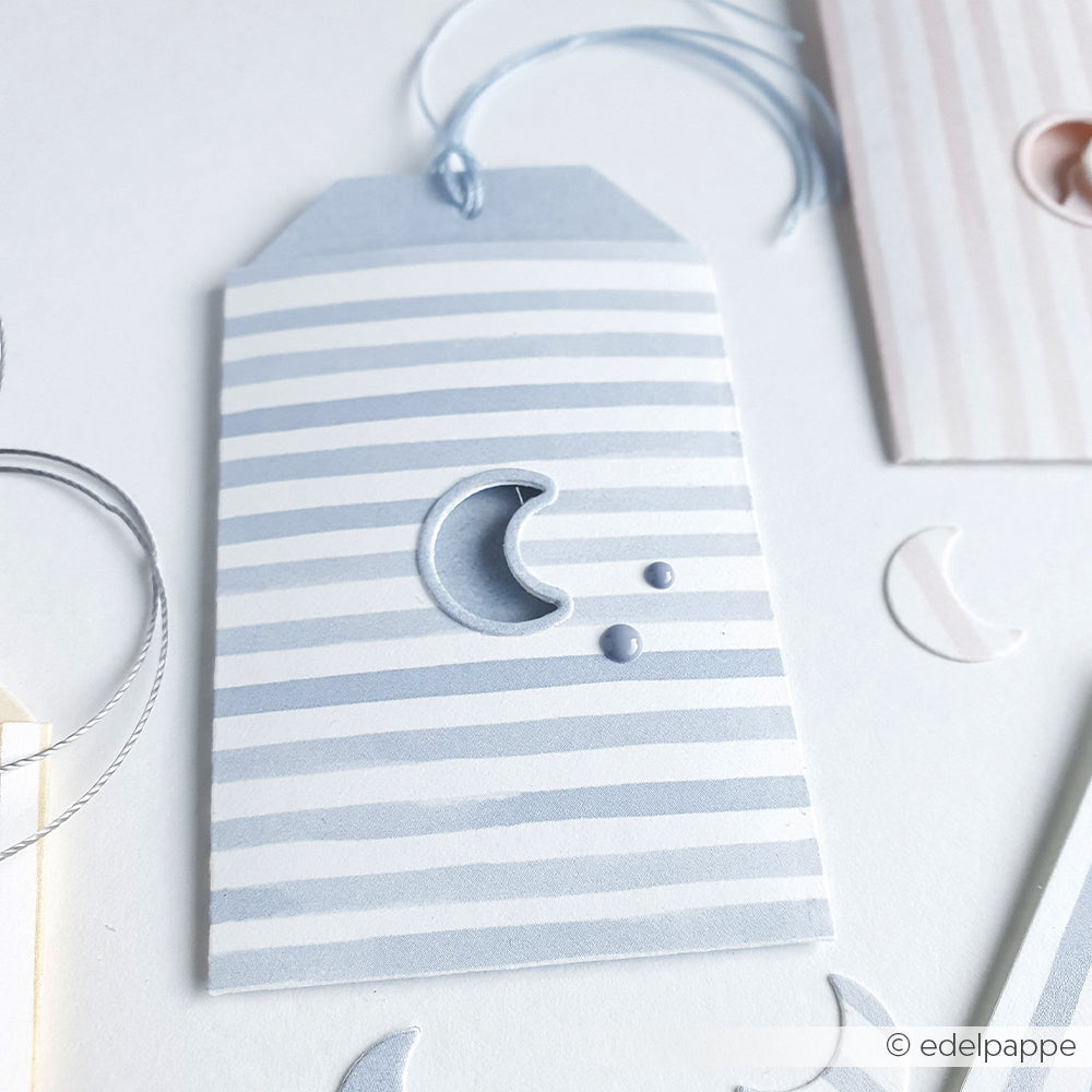 Der pastellblau gestreifte Geschenkanhänger von Alexandra Renke - Etikett im Tütchen-Mond - zeigt eine Mondsichel, die mit einer Motivstanze und zwei Punkten ausgeschnitten und mit einer blauen Kordel abgeschlossen ist.