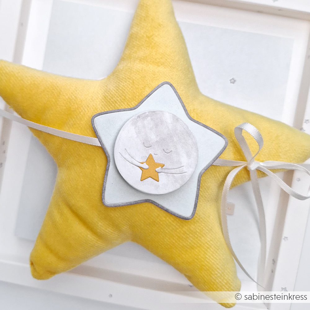 Gelbes Kissen in Sternform mit Schleife und Papierstern, auf dem ein verschlafenes Gesicht einen kleinen gelben Stern hält. Gestaltet mit Alexandra Renke Design Papers und dem Motivstanze Mond-Umarmung DieCut System für bezaubernde Details.