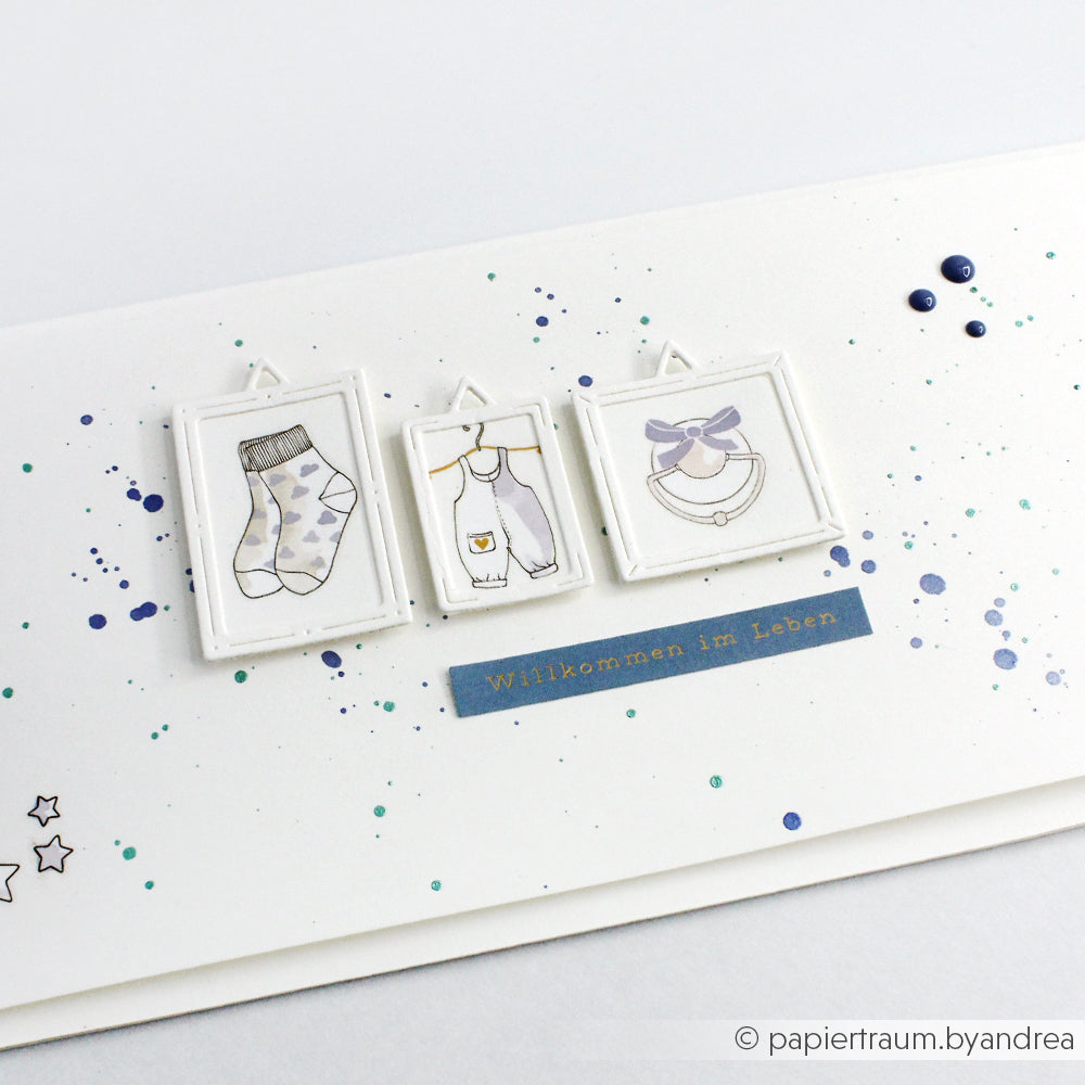 Eine handgefertigte Babykarte von Alexandra Renke mit gerahmten Zeichnungen von Socken, einem Strampler und einem Lätzchen mit Motivstanze Rechteckige Rähmchen, akzentuiert mit blauen und grünen Farbspritzern für zusätzlichen Charme.