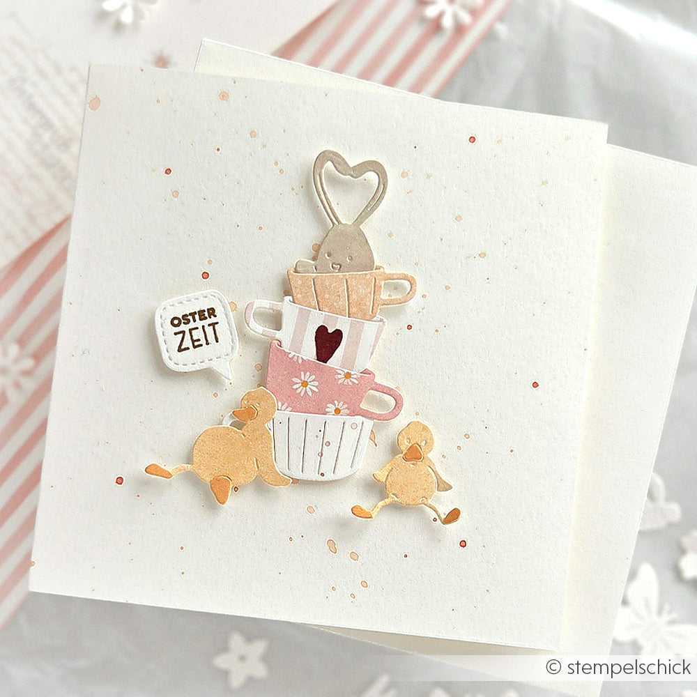 Handgemachte Karte mit gestapelten Teetassen aus der Motivstanze Fröhlicher Tassenstapel von Alexandra Renke, zwei gelben Küken und einem Schild mit der Aufschrift OSTER ZEIT. Hergestellt auf hochwertigem Designpapier für einen charmanten, festlichen Touch.