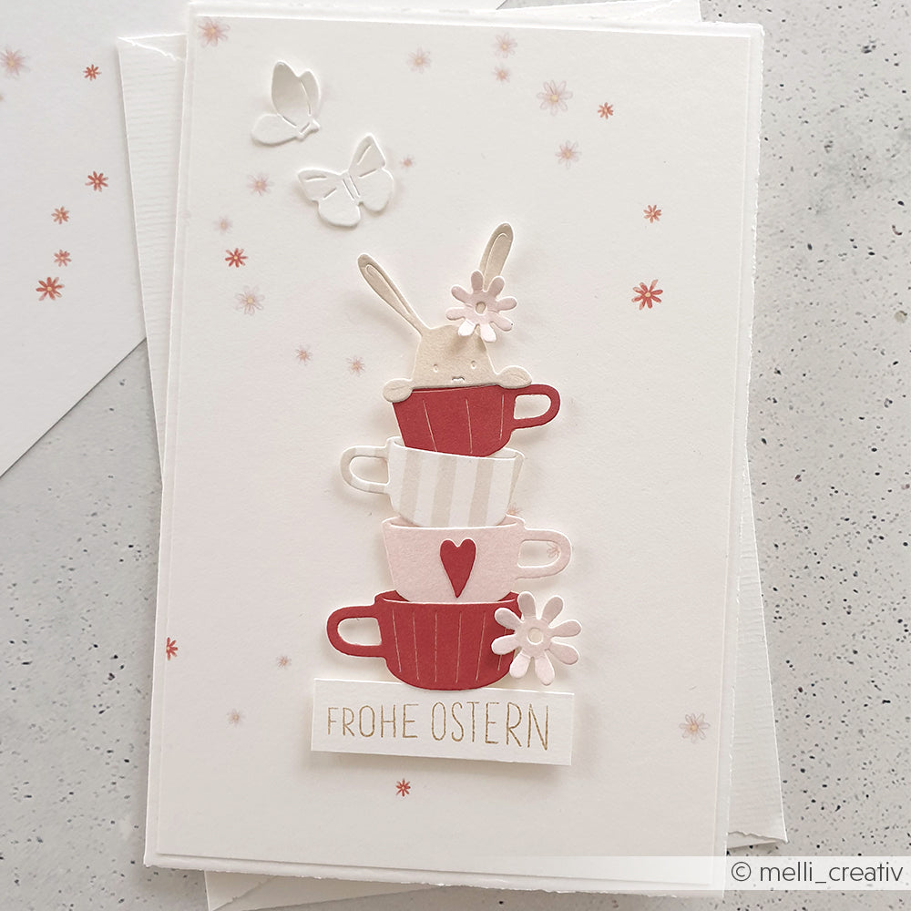 Alexandra Renke Grußkarte mit gestapelten Tassen, einem Hasen, Blumen, Schmetterlingen und "Frohe Ostern" in zarten Rosatönen - gestaltet mit Designpapieren und der Motivstanze Fröhlicher Tassenstapel für charmante Details.
