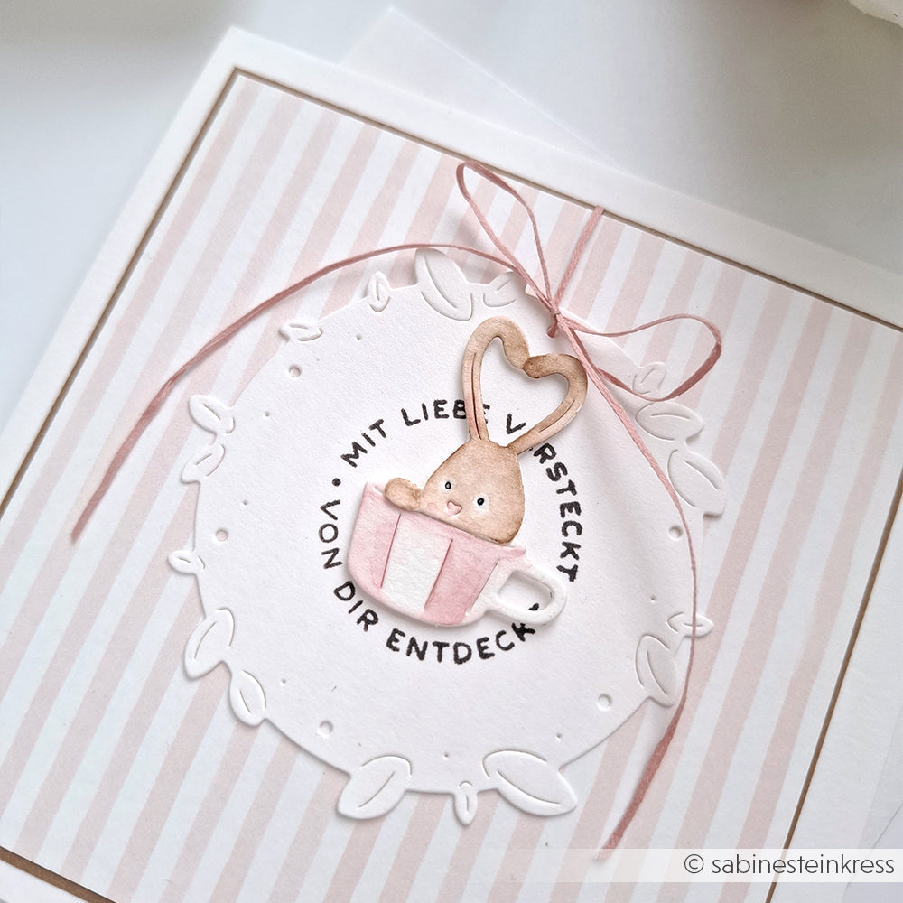 Ein niedlicher Hase in einer Teetasse mit Herz, umrahmt von Blättern und deutschem Text, auf einer gestreiften Karte mit rosa Schleife - perfekt zum Basteln mit Alexandra Renke Stanzsysteme oder Ihren Lieblingsmotivstanzen.