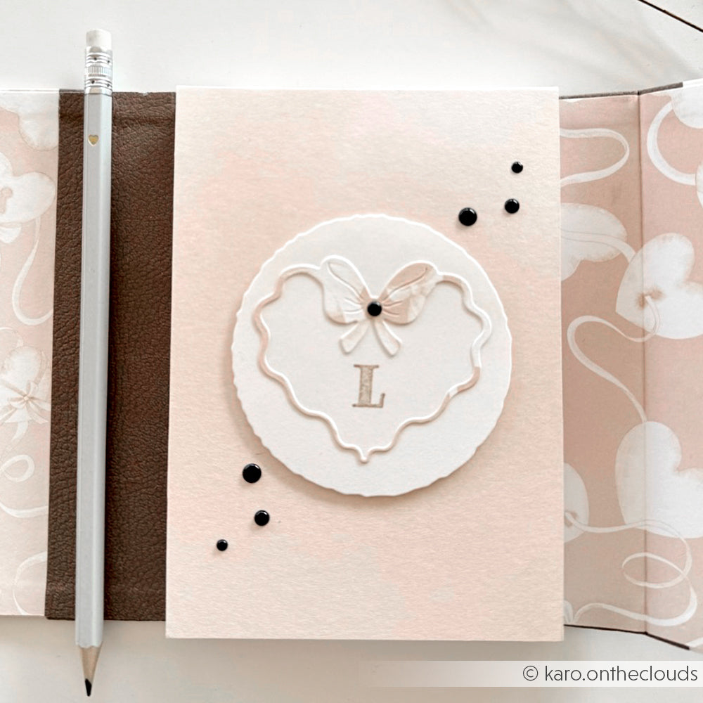Ein pastellrosa Alexandra Renke Notizbuch mit weißem Einband, Bleistift und schwarzem Perlendekor, monogrammiert mit 'L' und akzentuiert durch von Motivstanze Welliger Herz-Rahmen Stanzsysteme inspirierte Details.