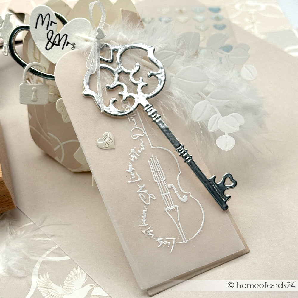 Eleganter Hochzeitsanhänger von Alexandra Renke, gefertigt aus hochwertigen Designpapieren. Mit Vintage-Schlüssel, weißen Federn, Geigenmotiv auf zartem Beige und filigranen Details von Motivstanze Schlüsselloch-Ornament.