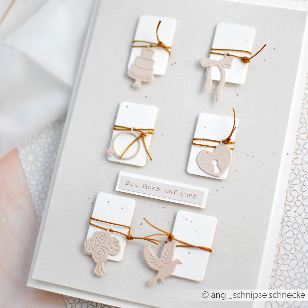Eine handgefertigte Karte von Alexandra Renke mit sechs weißen Anhängern mit goldener Schnur und einzigartigen beigen Anhängern, die präzise mit Stanzen für Designpapier hergestellt wurden, auf einem neutralen Hintergrund.