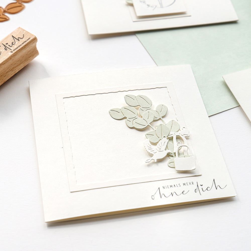 Eine handgefertigte Karte von Alexandra Renke mit Blättern und Vögeln, die mit DieCut Systemen und Stanzen für Designpapiere ausgeschnitten wurden, zeigt den deutschen Text "Niemals mehr ohne dich" auf einem sauberen weißen Hintergrund.