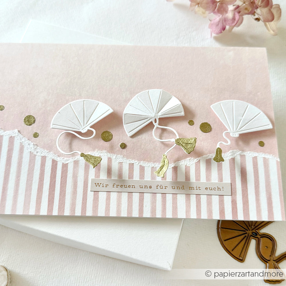 Handgefertigte Karte mit drei weißen Papierschirmen aus der Alexandra Renke Motivstanze Fächer Trio, mit goldenen Akzenten und deutschem Text auf einem rosa-weiß gestreiften Premium Designpapiere Hintergrund.