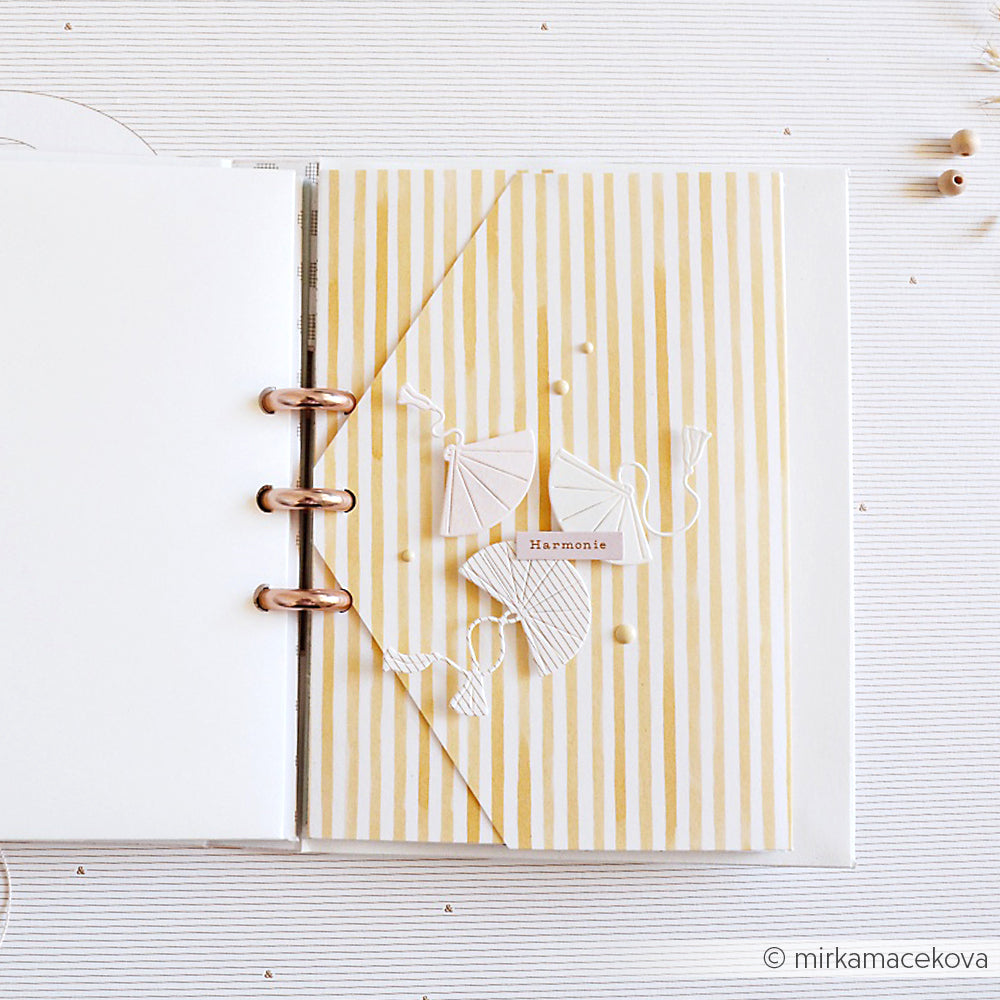 Offenes Scrapbook mit gelb gestreiften Alexandra Renke Designpapieren, weißen Drachenausschnitten mit einer Motivstanze Fächer Trio und "Harmonie" in der Mitte der Seite.