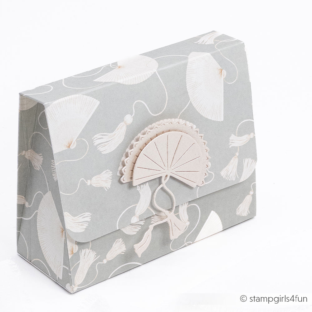 Eine dekorative Geschenktasche aus Papier von Alexandra Renke mit einem Fächer- und Quastenmuster in zartem Beige auf Hellgrau, hergestellt mit Designpapieren und DieCut Systemen für präzise Details und Eleganz.
