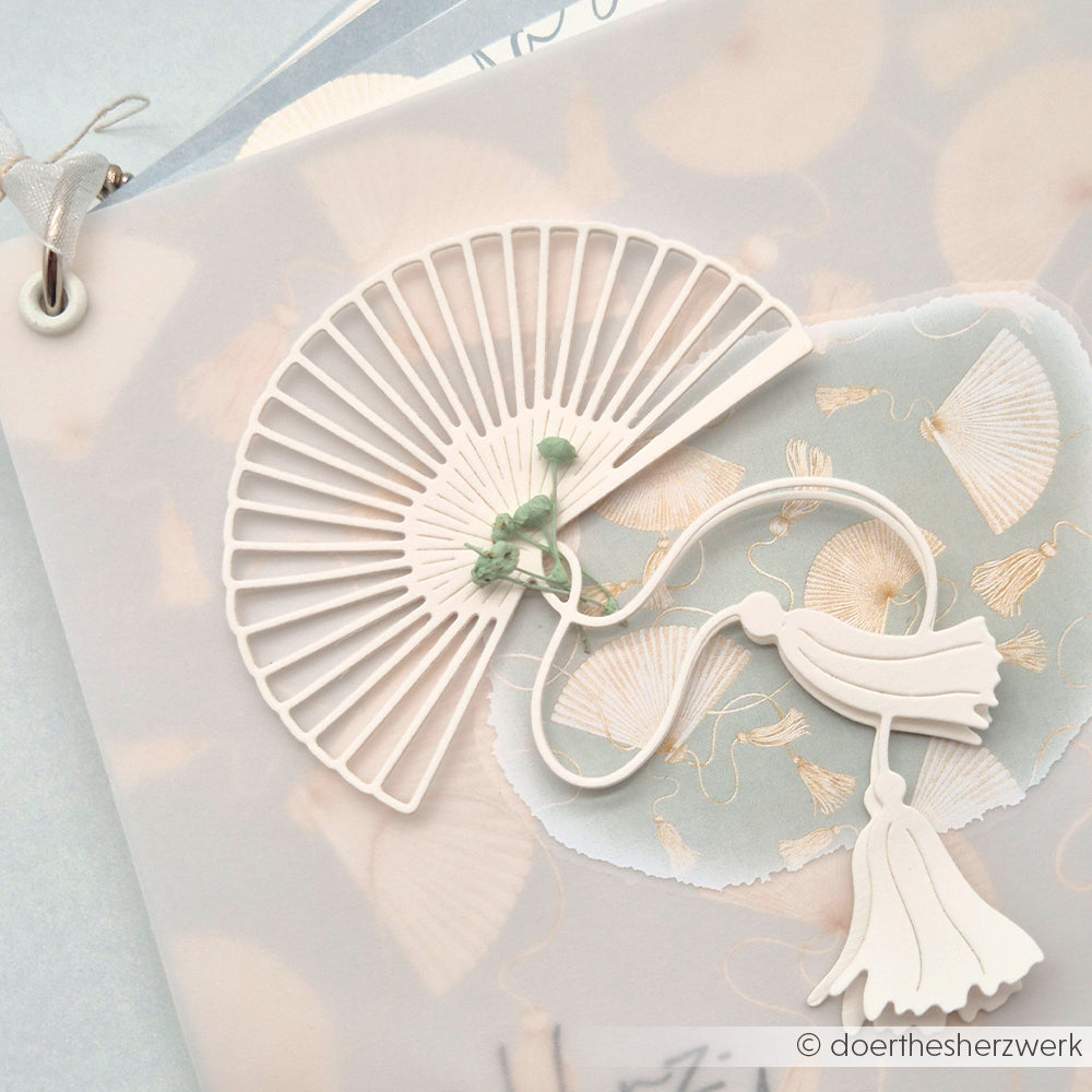 Zarter Papierfächer mit Alexandra Renke Designpapieren und der Motivstanze Großer Fächer, verziert mit Quasten, auf einem pastellfarbenen Hintergrund mit Muschelmuster.