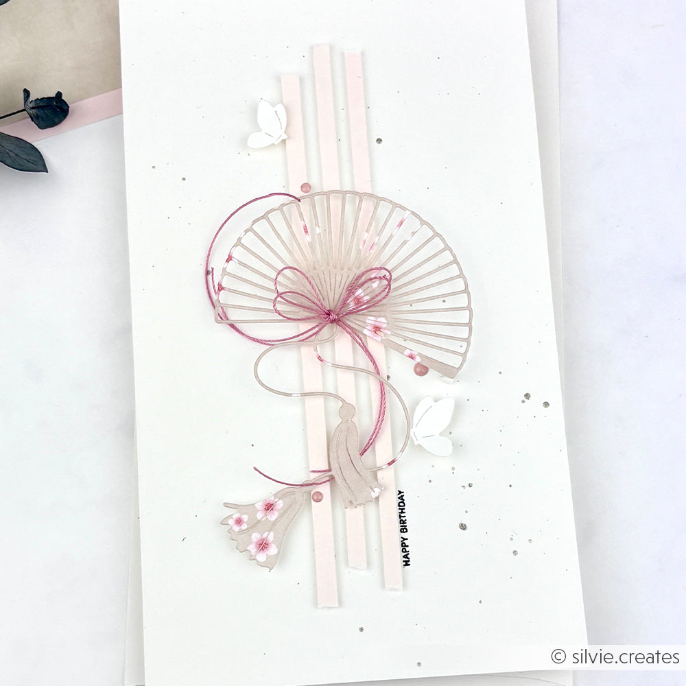 Elegante Geburtstagskarte von Alexandra Renke mit einem Papierfächer aus der Stanze Großer Fächer, verziert mit einer rosa Schleife, Blumen und Schmetterlingen auf weißem Hintergrund.