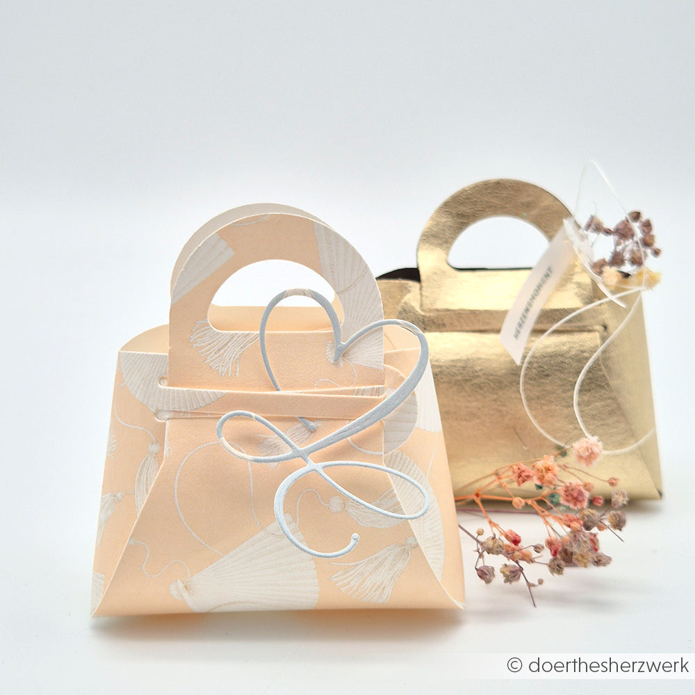 Zwei dekorative Papiertüten von Alexandra Renke - eine pfirsichfarbene und eine goldene - aus elegantem Designpapier werden mit getrockneten Blumen daneben auf einem hellen Hintergrund präsentiert.
