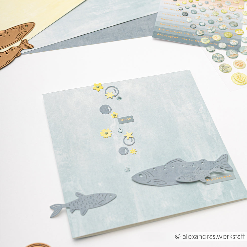 Scrapbook-Seite mit ausgeschnittenen Fischen, Seifenblasen aus der Alexandra Renke Motivstanze Luftblasen und Blumen auf blauem Hintergrund, dazu Alexandra Renke Designpapiere und Bastelmaterial.