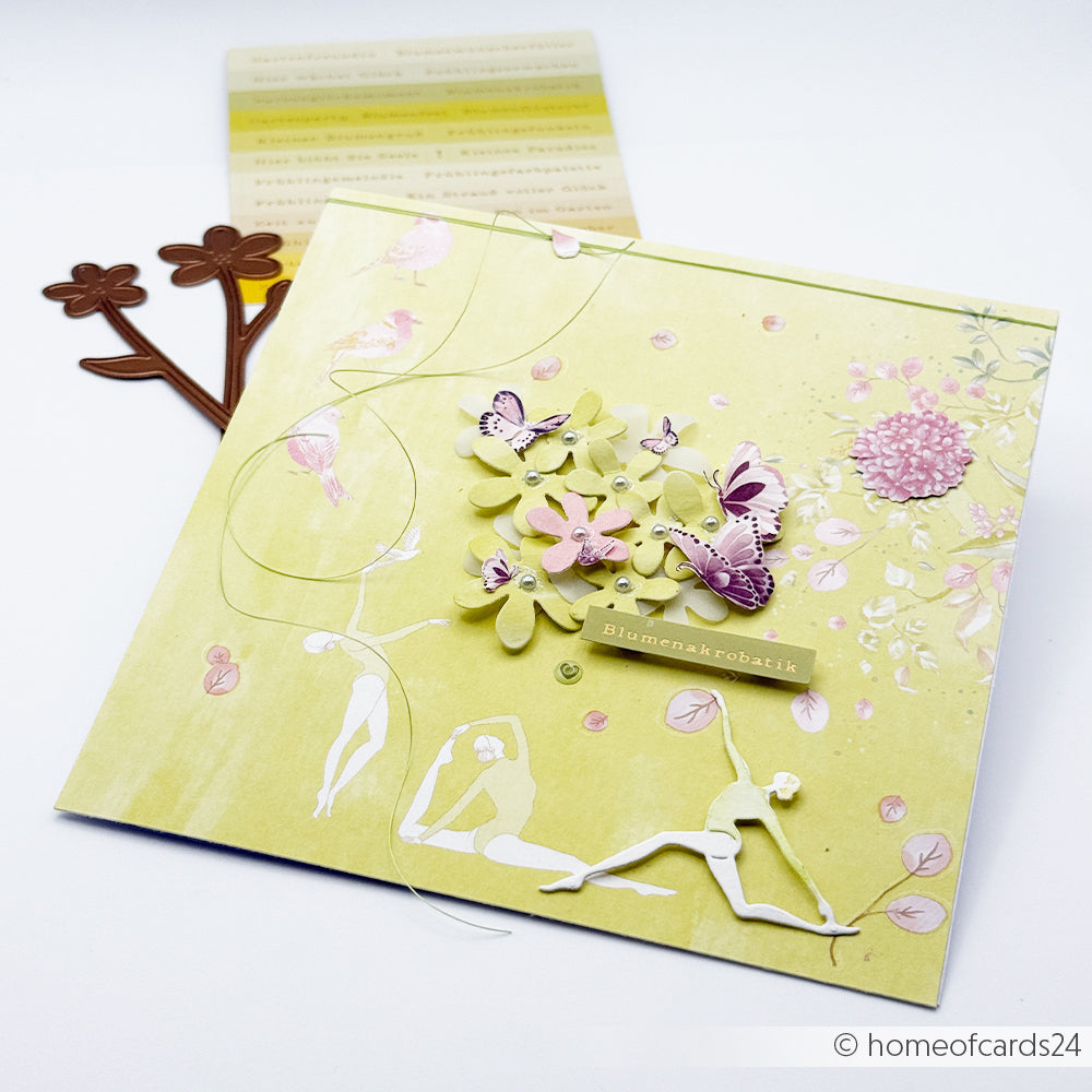 Handgefertigte Blumenkarte mit Schmetterlings- und Blumenausschnitten, gestaltet mit der Motivstanze von Alexandra Renke und Designpapieren, umrahmt von Bastelmaterialien auf weißem Hintergrund.
