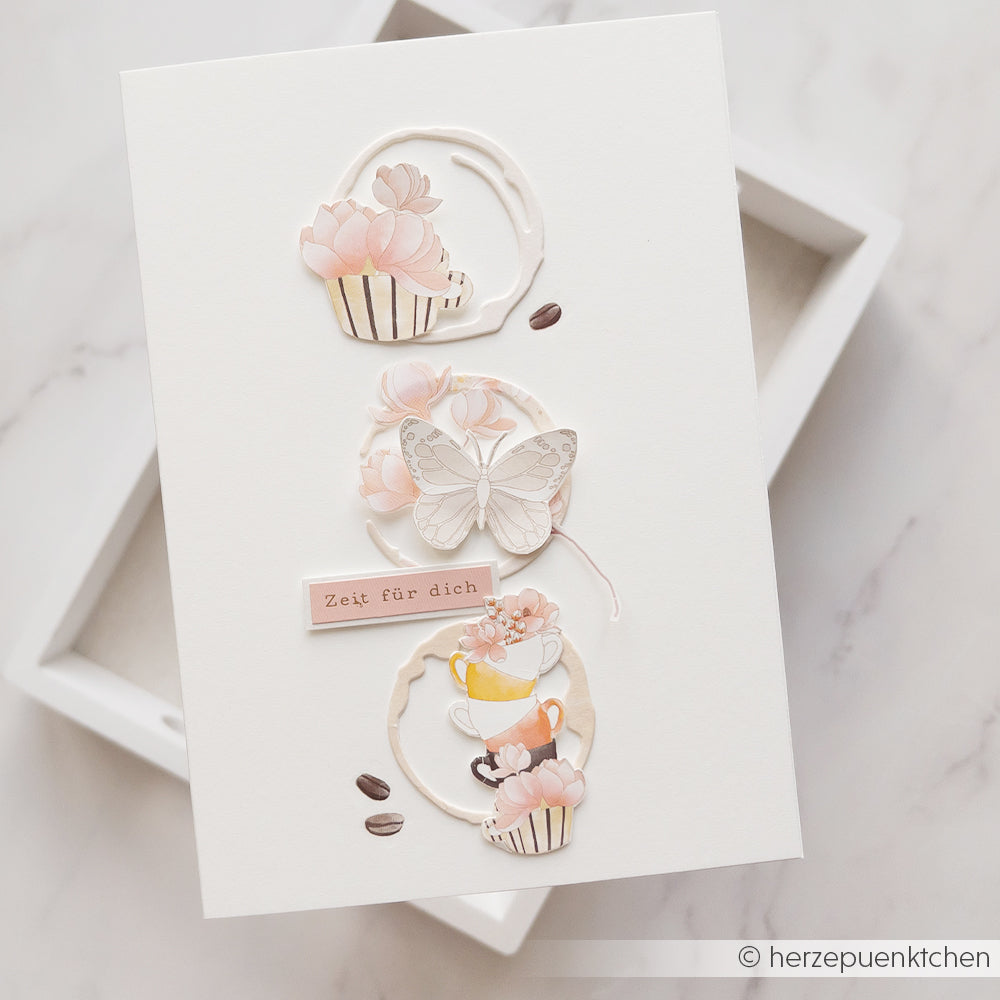 Handgefertigte Karte von Alexandra Renke mit pastellfarbenen Cupcakes, einem Schmetterling, Kaffeebohnen und dem Schriftzug „Zeit für dich“ auf weißem Hintergrund. Mit Kaffeering-Akzenten aus Designpapieren und der Motivstanze „Kaffeeringe“.
