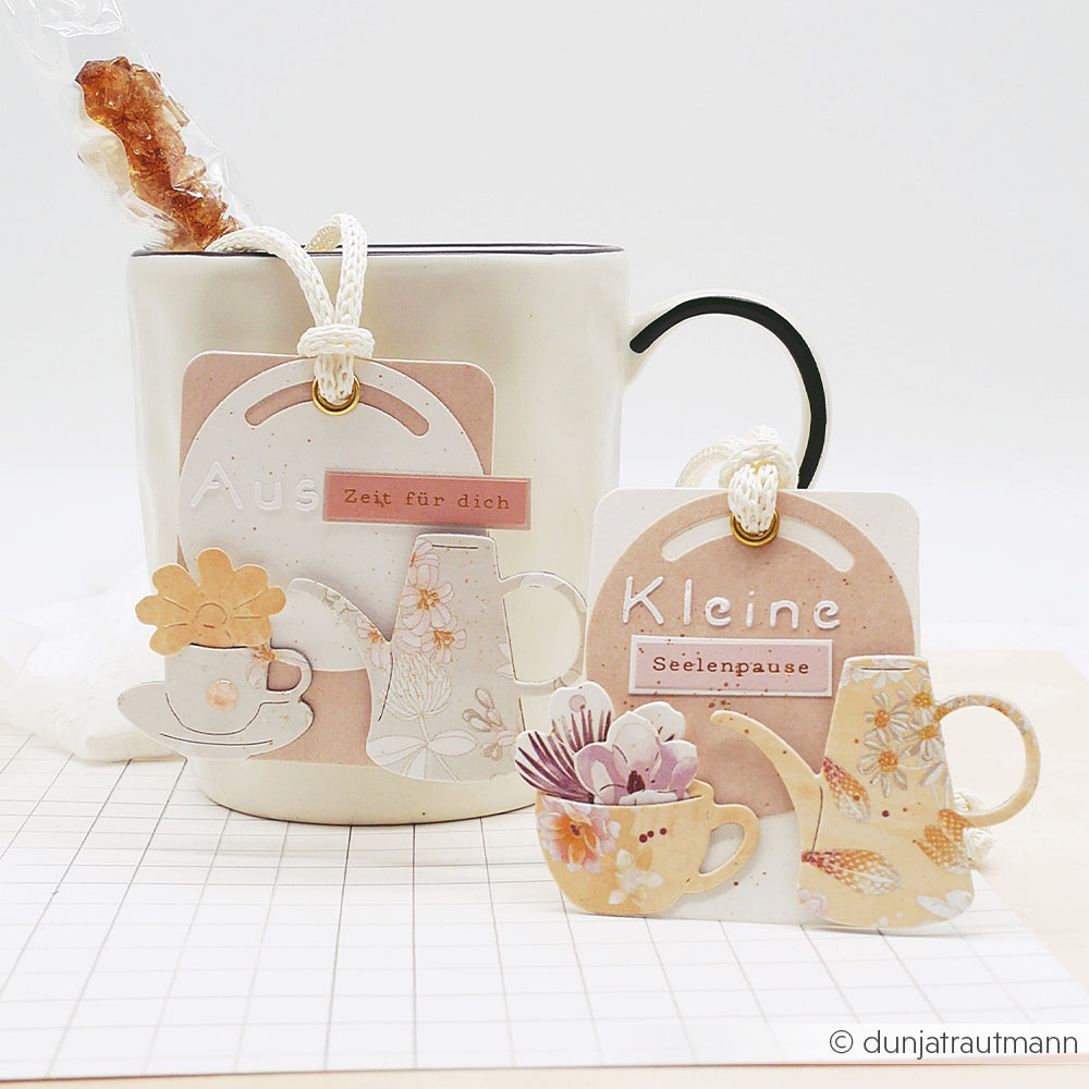 Weiße Tasse mit dekorativen Anhängern mit Teekannen- und Teetassenmotiven, gestaltet mit der „Motivstanze“ von Alexandra Renke und Stanzformen aus dem „blumigen Kaffeeset“, dazu ein Rührstäbchen aus Kandiszucker an der Seite.