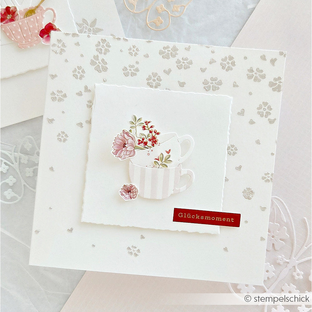 Weiße Karte mit geprägten Blumen, darauf zwei übereinander gestapelte Teetassen mit Blumenmotiven aus der Motivstanze „Blumiges Kaffeeset“ von Alexandra Renke sowie der Schriftzug „Glücksmoment“.