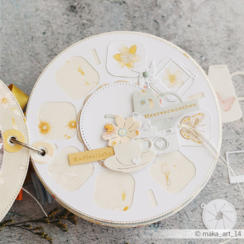 Eine runde Scrapbook-Seite mit Blumenmotiven, Ausschnitten der Kaffeetasse aus der „Blumiges Kaffeeset“-Motivstanze von Alexandra Renke sowie Verzierungen aus pastellfarbenem Designpapier, präsentiert auf einem grauen Hintergrund.