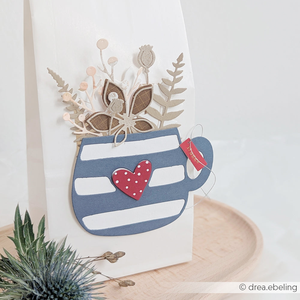 Geschenktüte mit einer blau gestreiften Tasse, inspiriert von der Motivstanze „Große Herztasse“, einem roten Herz und Papierblumen aus hochwertigen Designpapieren auf der Vorderseite, die auf einem Holztablett liegen.