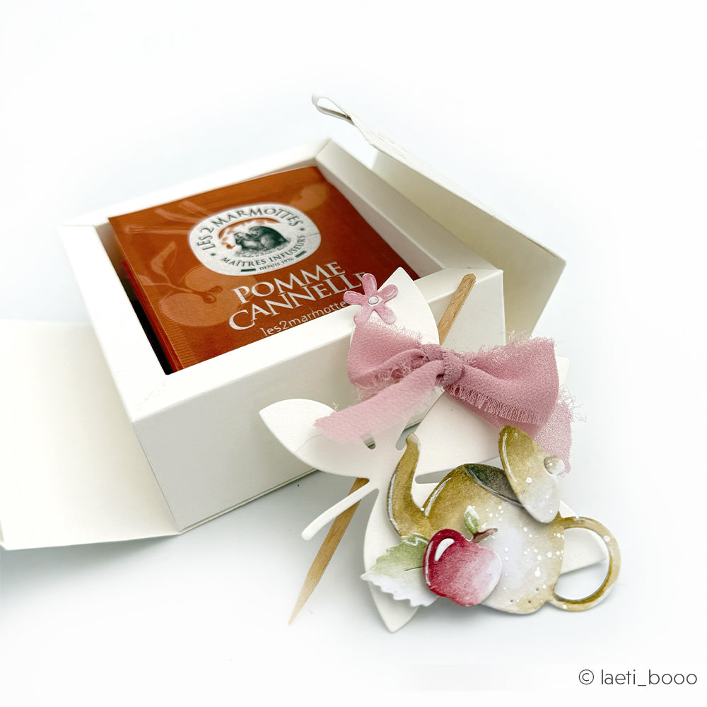 Teedose in weißer Verpackung mit einer rosa Schleife und apfelförmigen Verzierungen daneben, gestaltet mit der Motivstanze „Kleine Teekanne“ und den „Stanzen für Designpapier“ für ein bezauberndes, zartes Finish. Perfekt für Fans von DieCut-Systemen, die nach kreativer Inspiration suchen.