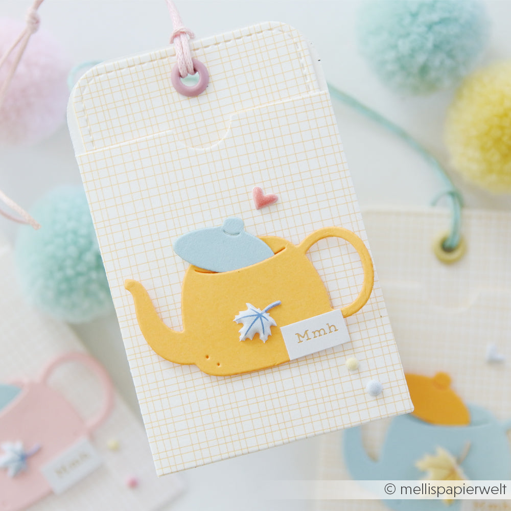 Gelbe Teekanne aus Papier mit blauem Deckel und einem Herz auf einem Anhänger, gestaltet mit der Motivstanze „Kleine Teekanne“ und dem DieCut-System, vor einem Rasterhintergrund und mit pastellfarbenen Verzierungen akzentuiert.