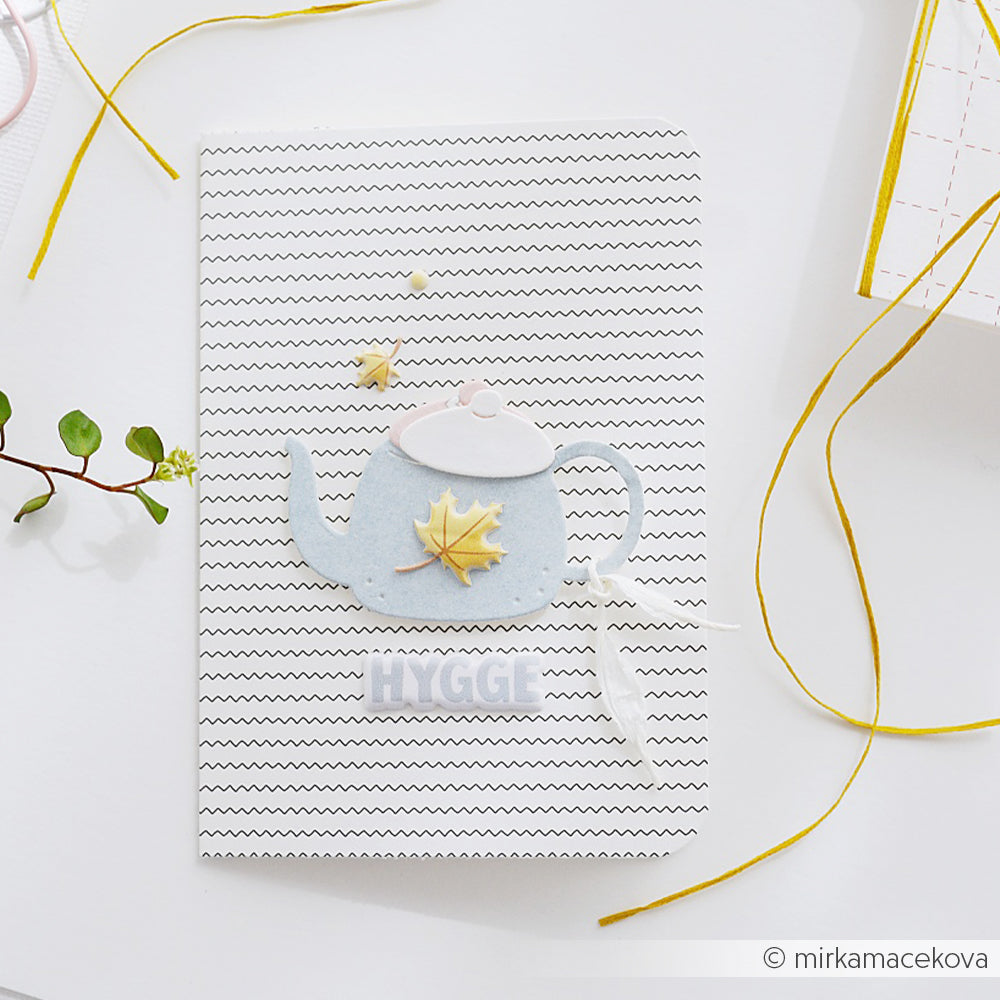 Eine Karte mit einer Teekanne, einem Blatt und dem Schriftzug „HYGGE“ auf weißem Zickzack-Hintergrund, wunderschön verziert mit gelber Schnur. Das bezaubernde Teekannen-Motiv wurde mit der Stanzform „Motivstanze Kleine Teekanne“ von „Qualitätsstanze und Stanzsysteme“ gestaltet und verleiht der Karte einen zusätzlichen Hauch von handwerklich inspirierter Gemütlichkeit.