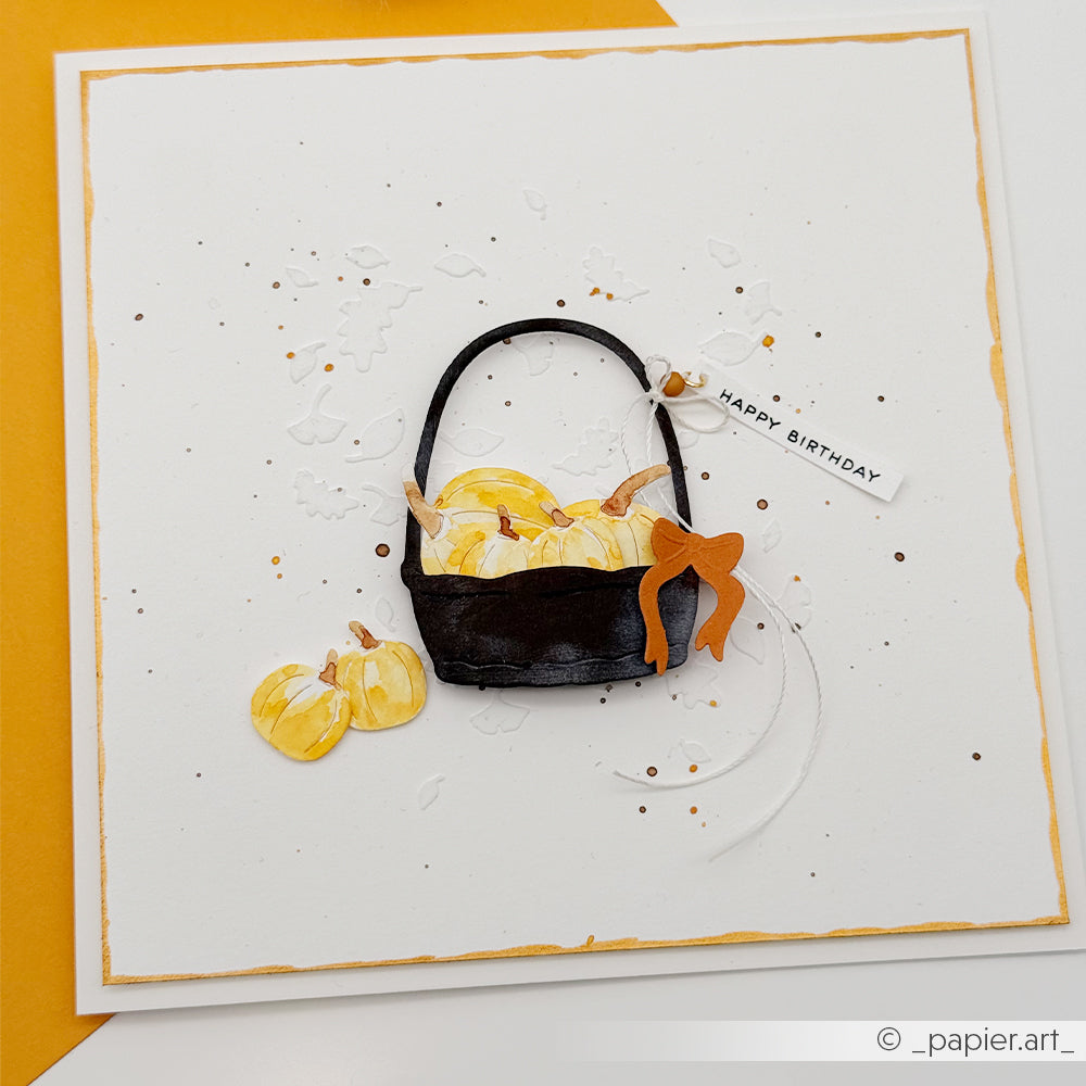 Handgefertigte Karte mit einem schwarzen Korb voller gelber Kürbisse, einer orangefarbenen Schleife und einem „Happy Birthday“-Anhänger auf weißem Papier, wunderschön gestaltet mit Stanzformen von Motivstanze und Stanzsystemen für präzise und elegante Details.