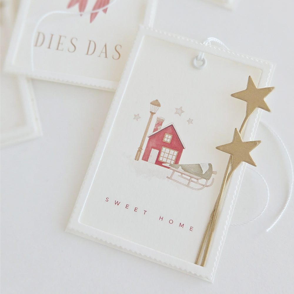 Ein Geschenkanhänger mit einer Aquarellillustration eines Hauses und eines Schlittens, verziert mit goldenen Sternchen aus dem „Sternenhimmel“-Set.