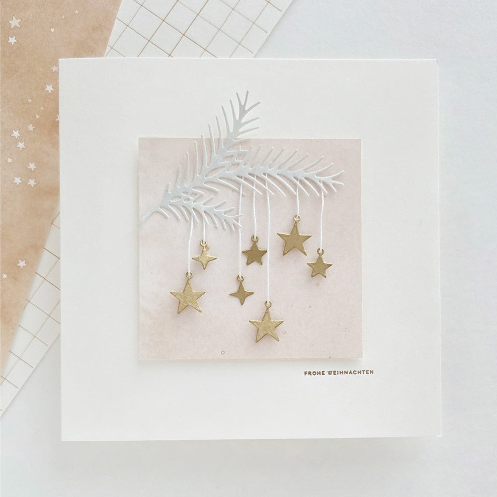 Eine quadratische Weihnachtskarte mit einem Tannenzweig aus Papier aus dem „Sternenhimmel“-Set, an dem kleine goldene Sterne hängen, sowie dem Schriftzug „FROHE WEIHNACHTEN“.