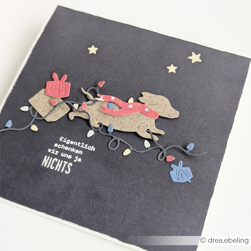 Eine handgefertigte Karte, gestaltet mit der Motivstanze „Lichterketten Duo“, auf der ein Dackel zu sehen ist, der sich in Weihnachtslichtern verheddert hat, mit dem deutschen Text: „Eigentlich schenken wir uns ja NICHTS…“.