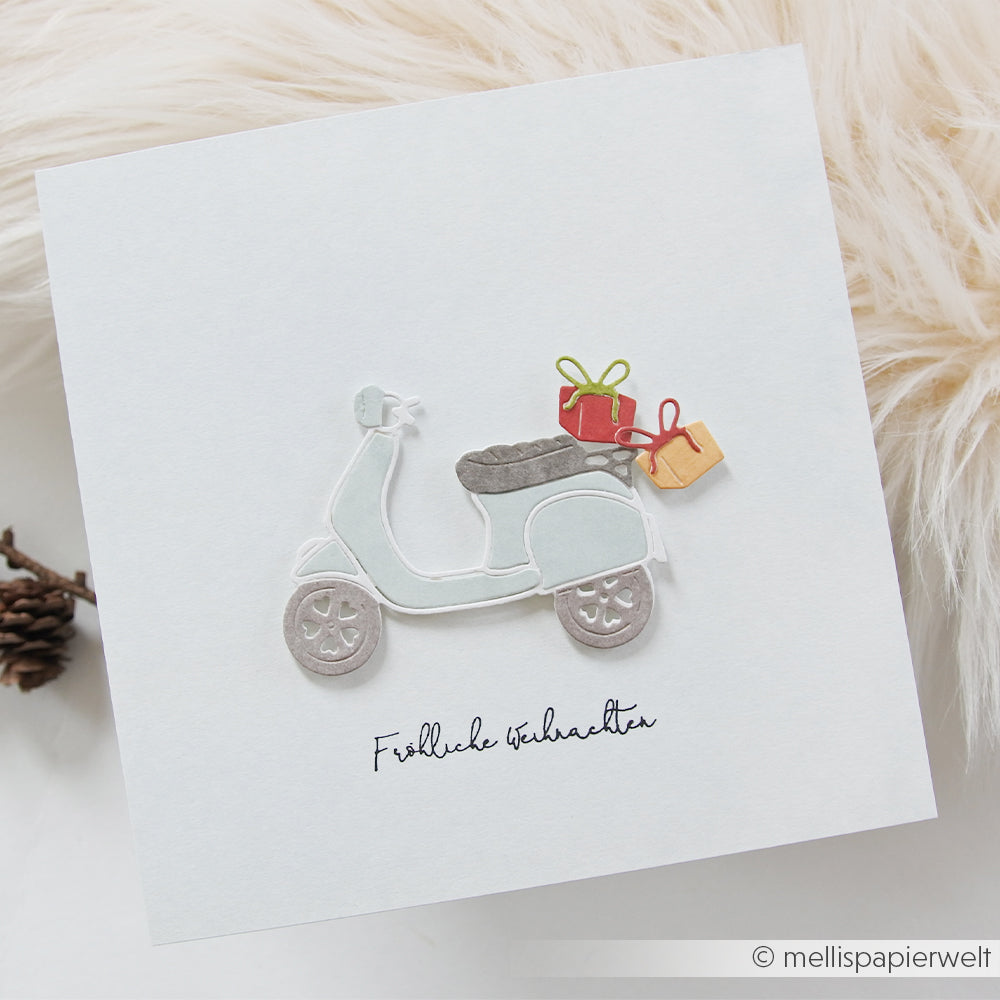Handgefertigte Grußkarte mit einem hellblauen „Geschenke-Roller“, der zwei Geschenkboxen transportiert, und dem Schriftzug „Fröhliche Weihnachten“.