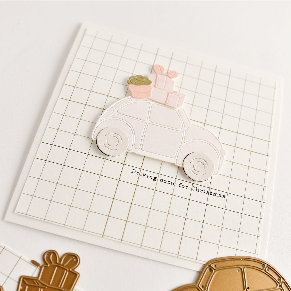 Eine Grußkarte auf kariertem Papier mit einem ausgestanzten „Geschenkeauto“, das mit Geschenken geschmückt ist, und dem Text „Driving home for Christmas“.