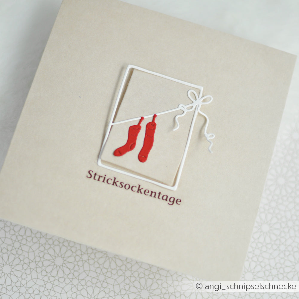 Grußkarte mit ausgestanztem Rahmen, zwei roten Socken an einer Schnur und dem Schriftzug Stricksockentage.