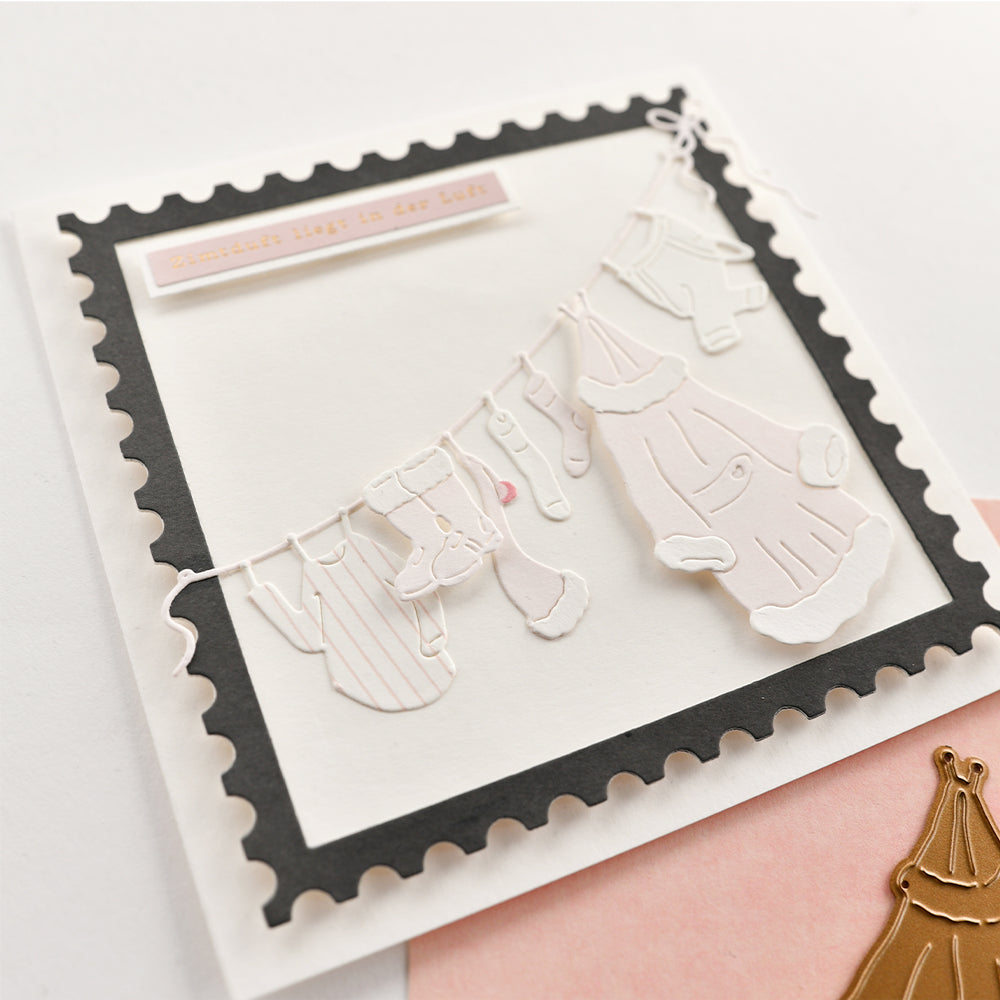 Eine handgefertigte Karte mit ausgestanzten Winterkleidungsstücken, die an einer Wäscheleine des Weihnachtsmanns innerhalb eines Briefmarkenrahmens hängen.