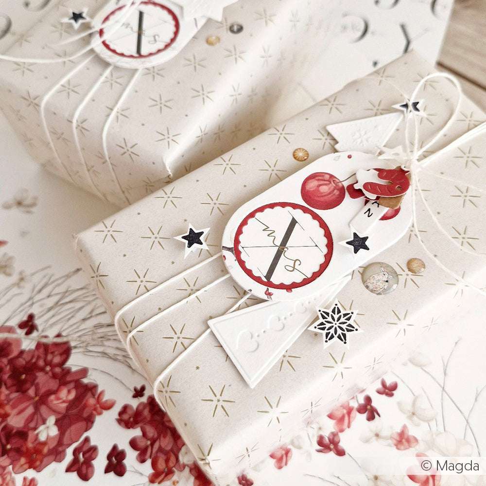 Zwei hübsch verpackte Geschenke mit sternförmigem Papier, weißer Schnur und festlichen Anhängern, die mit einem Stempel von Alexandra Renke hergestellt wurden, ruhen auf einem floralen Hintergrund.