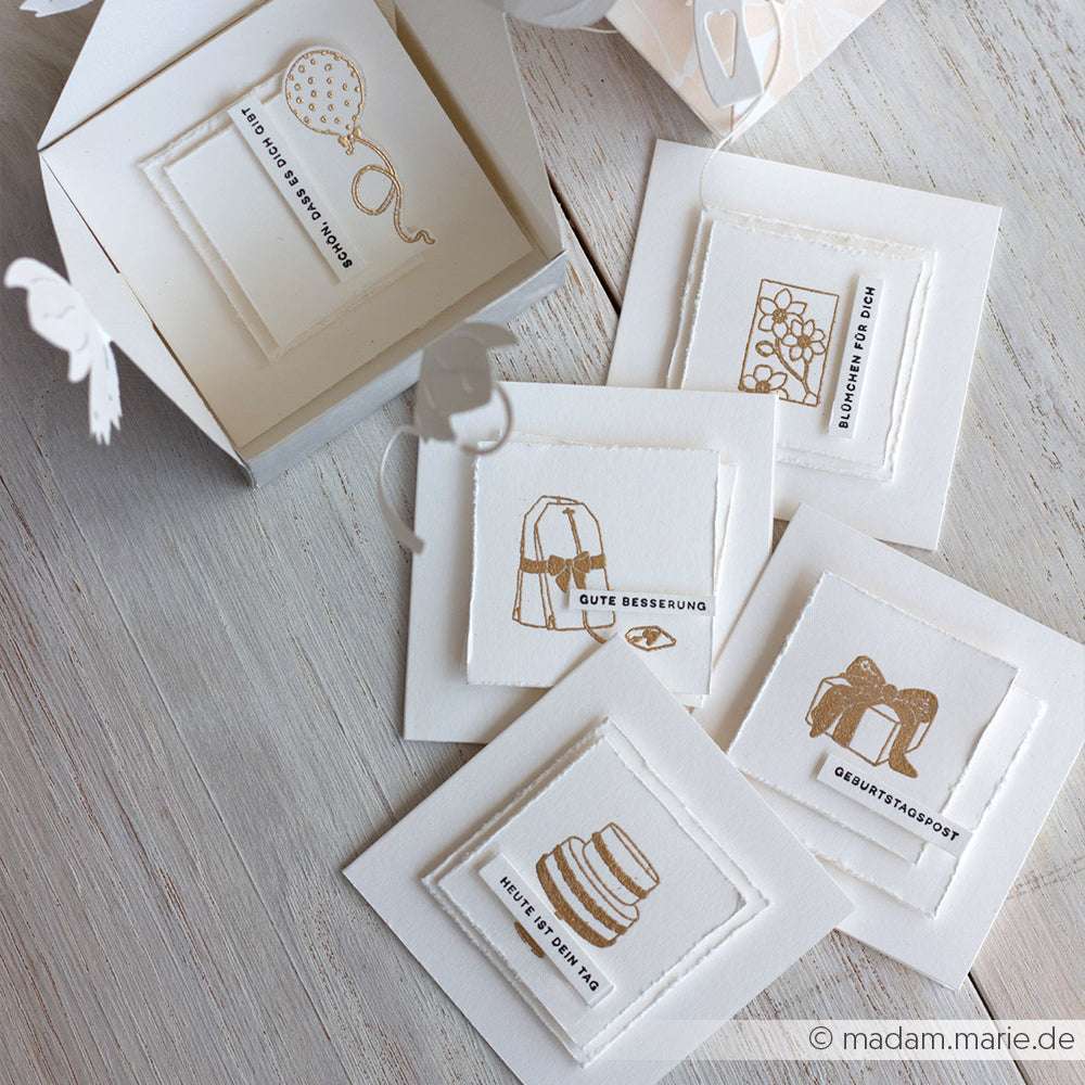 Vier weiße Alexandra Renke-Grußkarten mit goldenen Linien und deutschem Text, arrangiert in einer offenen weißen Geschenkbox - ideal für Stempel für Papierprojekte oder um einen Teebeutel voller Liebe zu Ihrem nächsten Projekt hinzuzufügen.
