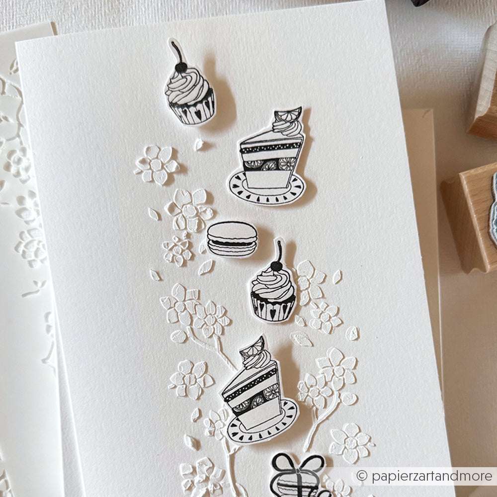 Weiße Karte von Alexandra Renke mit geprägten Blumen, gekrönt von schwarz-weiß ausgeschnittenen Torten, Cupcakes, einem Macaron und einem Kirsch-Cupcake-Motiv - ideal zum Basteln mit Ihren Lieblingsmotiven von Holzstempel oder Stempelgummi.