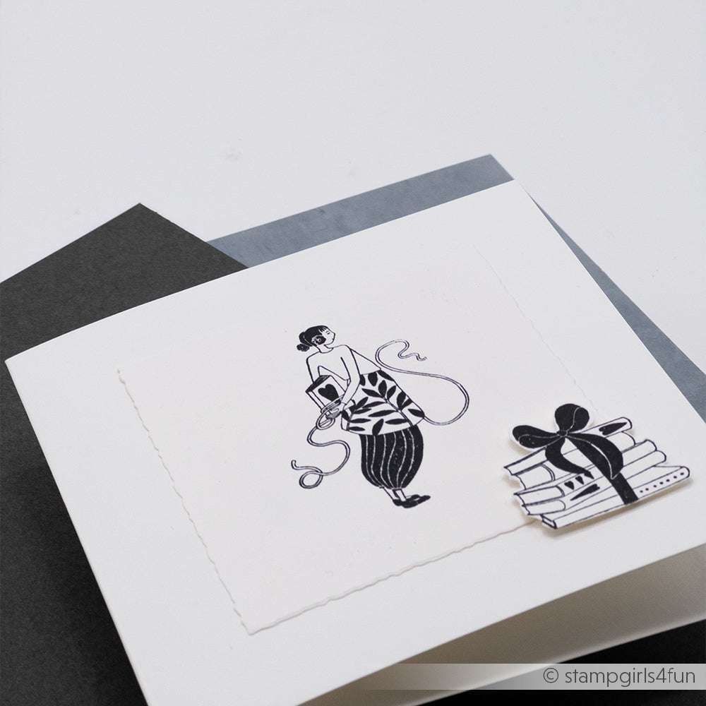 Eine handgefertigte Karte von Alexandra Renke mit einer Frau, die Garn in der Hand hält, und einem Stapel Bücher, der mit einem Band verschnürt ist - perfekt für Bücherliebhaber.