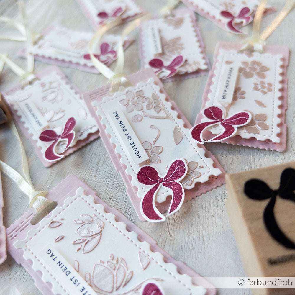 Auf einer hellen Fläche sind mehrere rosa-weiße, handgefertigte Geschenkanhänger mit Blumen- und Schleifenmotiven arrangiert, die mit hochwertigen Holz- und eleganten Holzstempeln von Alexandra Renke wunderschön akzentuiert sind.