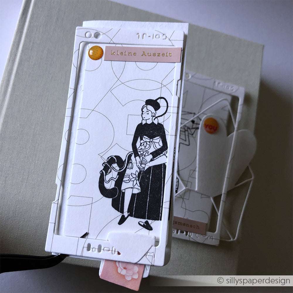 Eine Schwarz-Weiß-Illustration einer Frau und eines Kindes erscheint auf einer mehrschichtigen, dekorativen Papierkarte über einem geschlossenen Buch mit Akzenten, die von Alexandra Renke-Designs inspiriert sind.