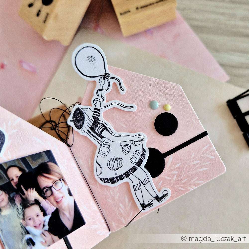 Alexandra Renke Papierkarte in Form eines Hauses mit einem schwarz-weißen Ausschnitt, Figurinen Kind mit Luftballon, neben einem Familienfoto.