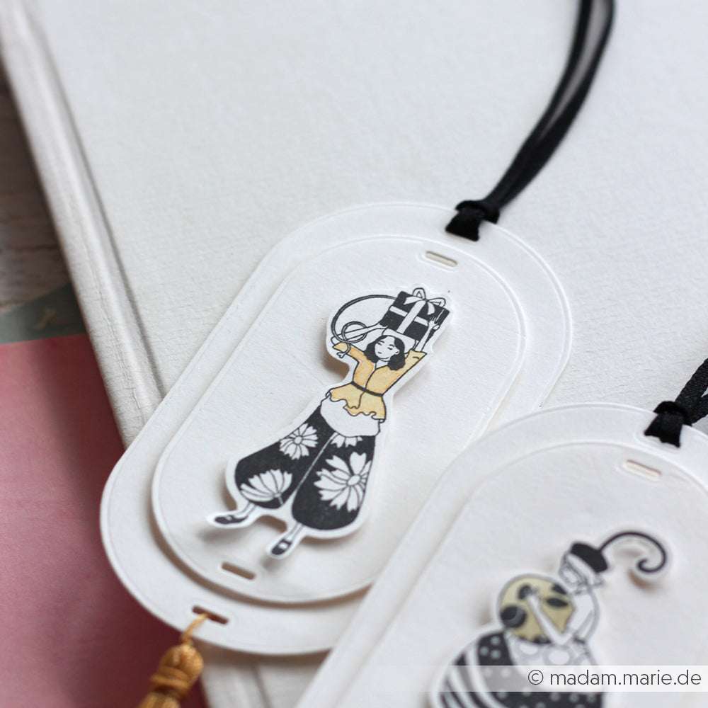 Ein Papierlesezeichen im Vintage-Stil von Alexandra Renke mit einer illustrierten Frau und einem schwarzen Band, das auf einer weißen Oberfläche ruht - ideal für Stempeln für Anfänger oder als Ergänzung zu Ihren Lieblings-Holzstempel-Designs.