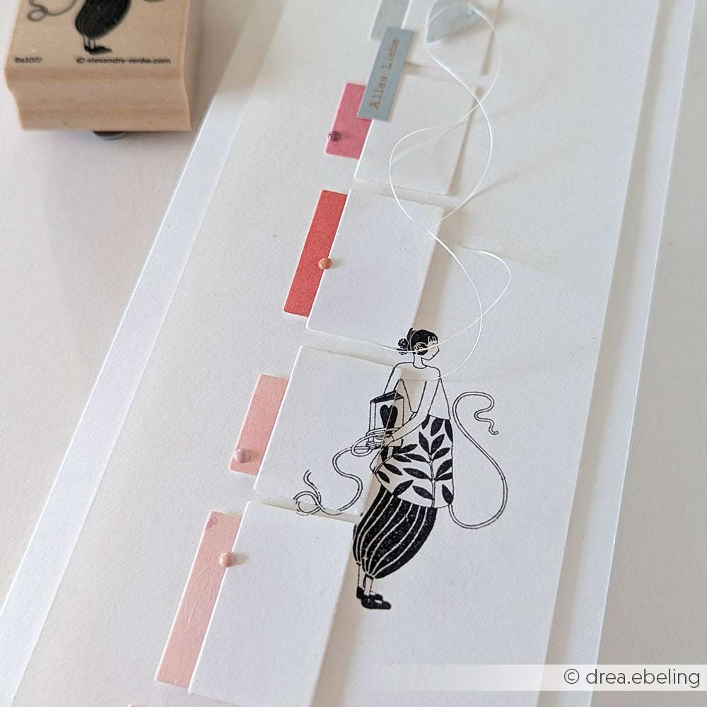 Alexandra Renkes Kunstdruck einer Frau mit rosa und roten Anhängern, die an einer Schnur befestigt sind, ruht auf einer weißen Fläche neben einem Holzstempel.